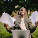 Super positive black student sitting in green park with application papers and laptop and smiling happily after learning news about being accepted to university