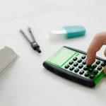 Close-up of a person's hand pressing a calculator on a desk with other stationery items.