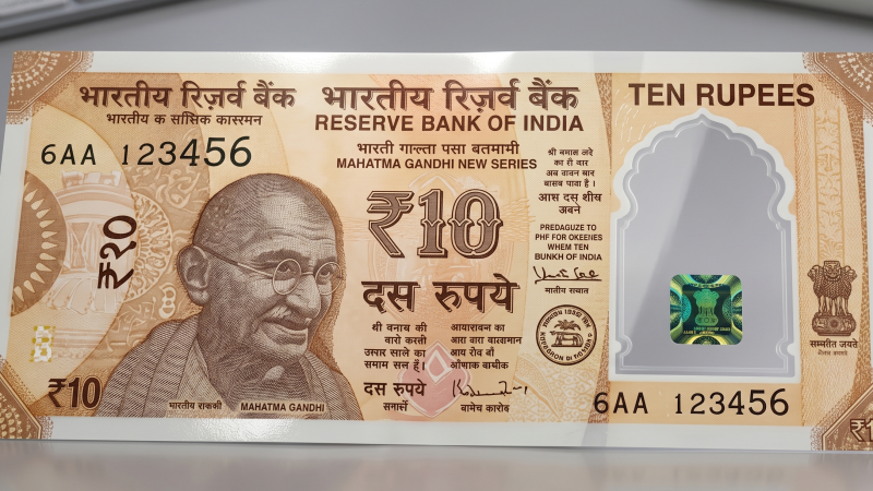 A realistic concept design of the Reserve Bank of India (RBI) new ₹10 polymer plastic currency note, featuring Mahatma Gandhi and a prominent translucent, transparent security window with a holographic emblem, displayed flat on a clean, modern grey office desk.